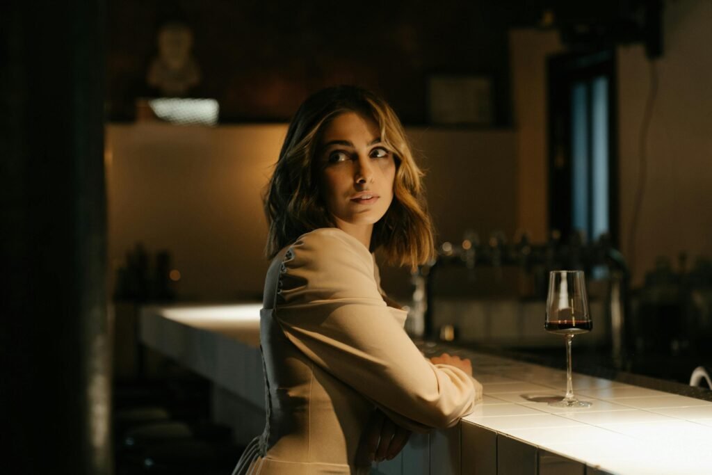 A glamorous woman in a stylish dress sits at a bar counter, elegantly sipping wine.